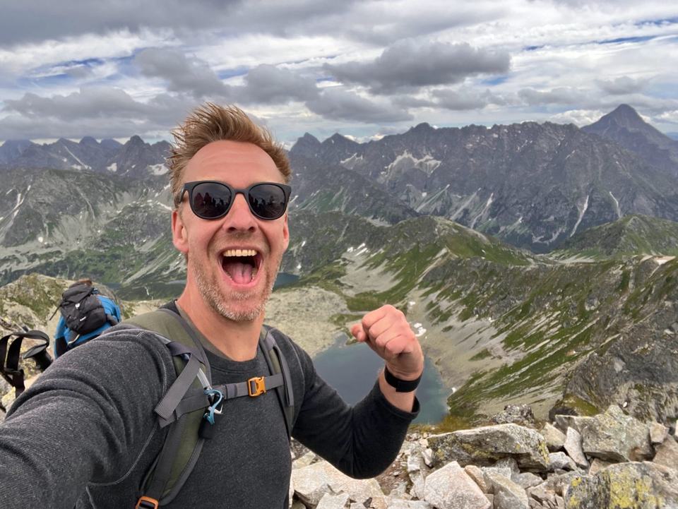 Sierk Rijkeboer juicht op een bergtop, met achter hem een bergmeer en scherpe pieken